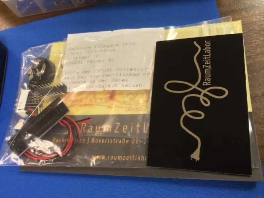 A plastic bag with an electronics kit with microcontroller and display, some RaumZeitLabor merchandise and an obituary notice. A plastic bag with an electronics kit with microcontroller and display, some RaumZeitLabor merchandise and an obituary notice.