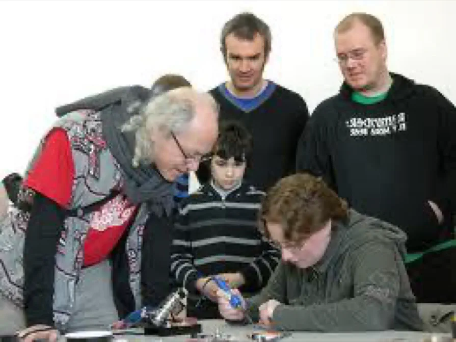 Mitch Altman helping a participant solder, surrounded by onlookers Mitch Altman helping a participant solder, surrounded by onlookers