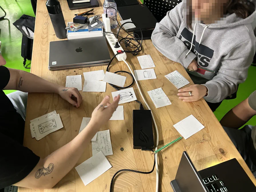 Nach den Crazy 8s werden die Ideen geclustert Skizzenblätter liegen auf einem Tisch verteilt, zwei Personen ordnen und besprechen sie