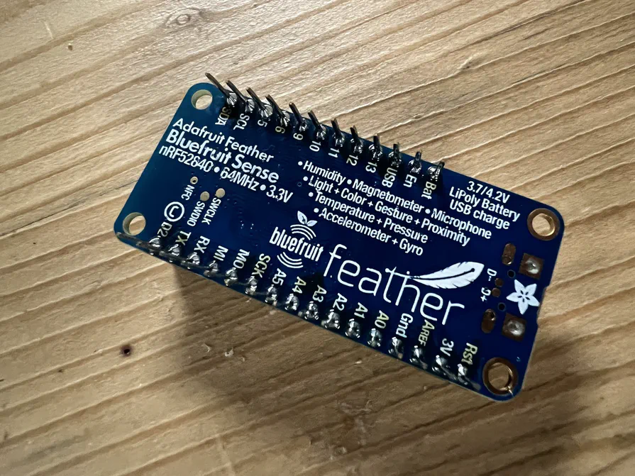 Das Board, das durch den Kurs führte: der Adafruit Feather nRF52840 Sense Ein Adafruit Feather nRF52840 Sense auf einem Holztisch, die Rückseite mit der Liste der eingebauten Sensoren nach oben