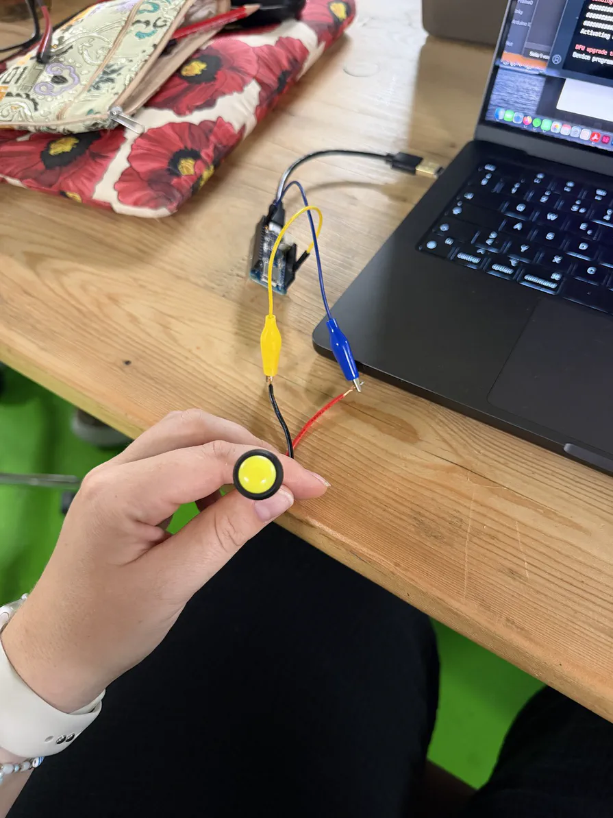 Ein Taster, zwei Krokodilklemmen und ein Board: fertig ist die eigene Tastatur Eine Hand hält einen gelben Taster, der über Krokodilklemmen mit dem Board neben einem Laptop verbunden ist